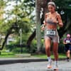 Einen Halbmarathon laufen weltweit über 2 Millionen Menschen pro Jahr. Eine Frau in einem beigefarbenen Sportoutfit mit Startnummer läuft auf der nassen Straße.